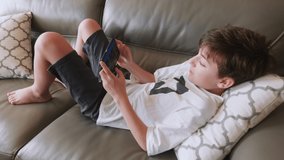 Happy boy plays emotionally game on smartphone is lying on sofa. Child plays bedtime. Internet. Gamer. Quarantine. Online education. Lifestyle. Hobby. Relax. Childhood