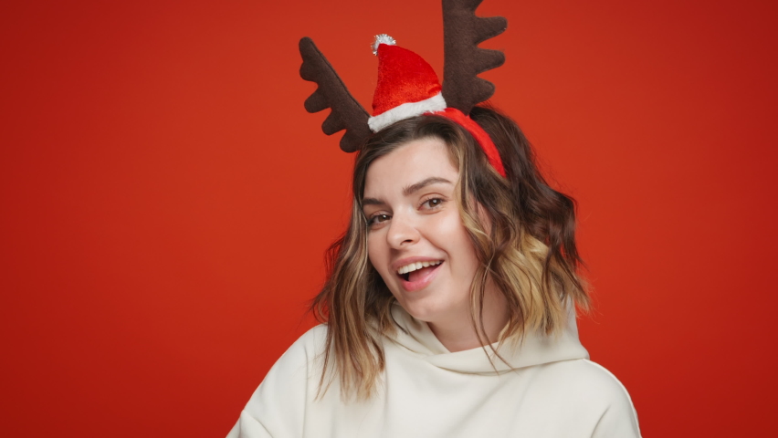 Happy young woman in Christmas clothes hat Santa dancing sings to music rhythmically moving her fingers with Candy deer horns  smiling happily looking at camera on red background. Happy New Year. Noel