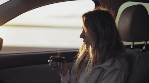 Woman inside the car is drinking coffee from the thermos cup, adoring sunrise