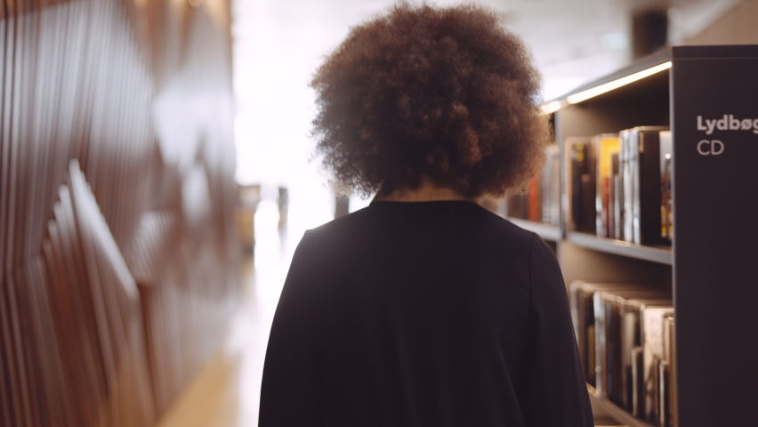 Tracking Medium Handheld Rear View Shot Of Young Woman With Afro Hair In Black Walking Between Bookshelves Of Library