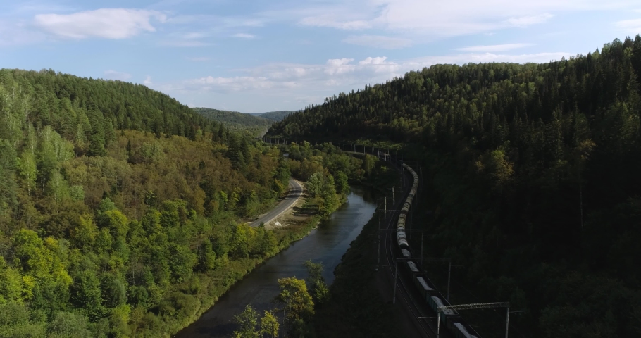Freight long train carries with oil tank and petrol carriages an electric locomotive by Trans Siberian railways under the rock and near mountain river. Aerial drone wide view at summer sunny day Royalty-Free Stock Footage #1080832238