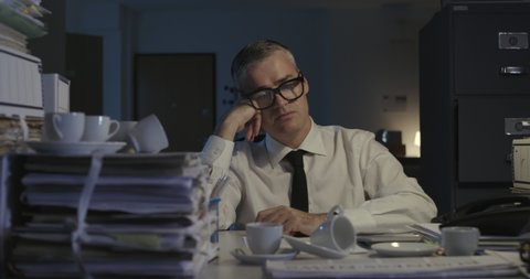 Stressed tired corporate businessman working overtime in his office, he is surrounded by piles of paperwork