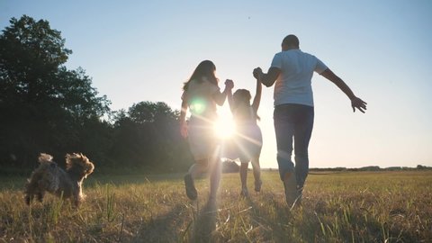Happy family in park at sunset with pet. family has fun with child. Parents hold child by hands. Helping hand of parents. Walking happy family with pet in park at sunset.