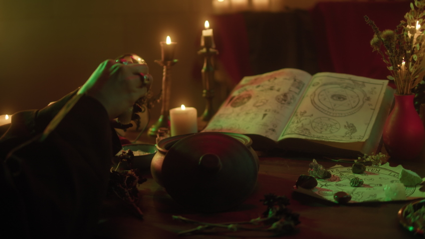 Witch cooking potion elixir close-up. Table with spellbook for making rite. Herbs, burning candles, crystal magic ball for sacrifice. Halloween theme, full moon, witchcraft. 