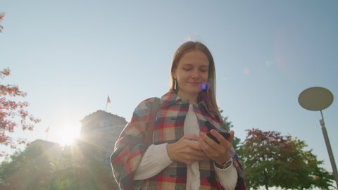 Confident millennial Lady uses by Smartphone App outside near Bundestag in Berlin in Sunny Autumn Day. Happy Businesswoman in Coat in German Capital. Medium Long Low Angle 4K Slow Motion orbit Shot