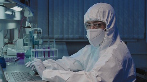 Tilt up slowmo of unrecognizable female scientist in coveralls, face mask, gloves and goggles working on laptop in laboratory, then looking at camera