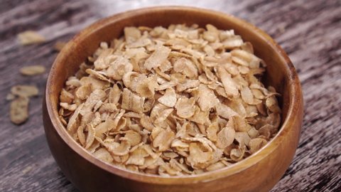 Uncooked dry cereal spelt wheat flakes falling into rough wooden rustic bowl. Close up. Rotation. On a wood surface. Healthy superfood concept. Slow motion