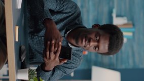 Vertical video: Black man in living room enjoying social media content on phone, smiling while using modern wireless technology. African american guy relaxing at home, online internet activity