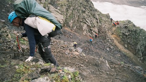 The climber climbs up the mountain with a backpack with equipment, fastened with a carabiner with a belay to the rope. A man climbs up to the top of the mountain