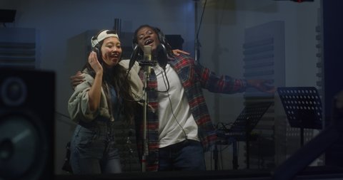 Cinematic shot of young professional smiling energetic band singers wearing headphones are performing together a new song with a microphone while recording it in a music studio with manager.