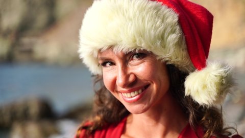 Portrait of a woman with makeup smiling at the camera and playing in a Santa hat on the beach, seashore. 4K