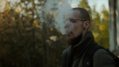 A bearded man in glasses smoking electronic cigarette blowing smoke and enjoying it. A vaping person with backpack standing outside looks like getting high. Blurred trees background
