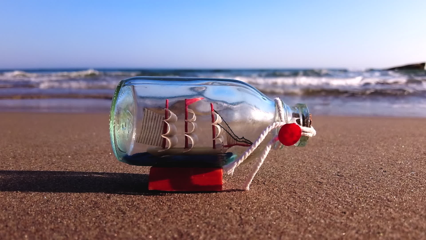 A ship in a bottle lies on a sandy beach and is washed by waves in slow motion. 