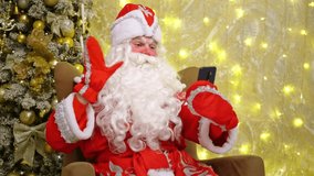 Happy old bearded santa claus in suit waving his hand holding cell phone, using mobile application for video calls, greeting merry christmas on smartphone in virtual online chat, christmas tree