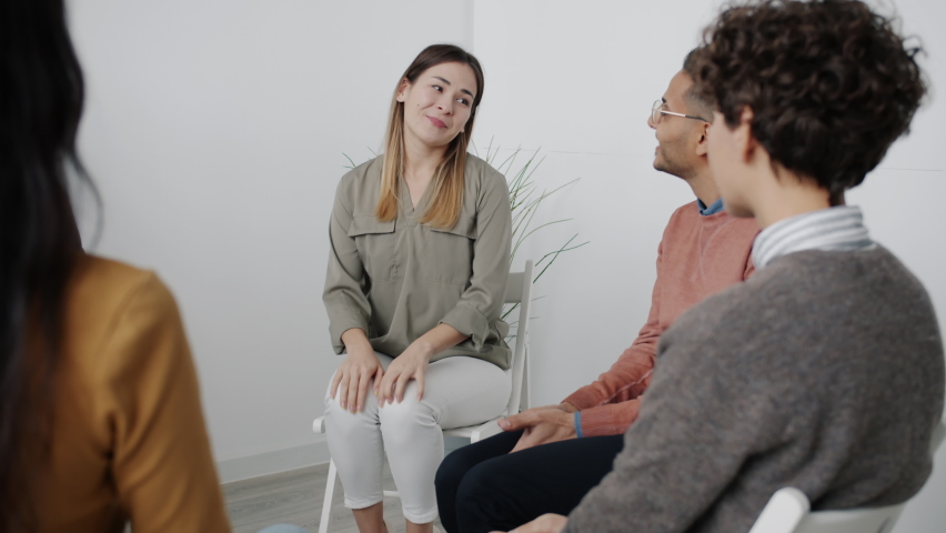 Nervous young woman is sharing feelings during group therapy sitting in circle in white room talking and gesturing while people expressing support and understanding