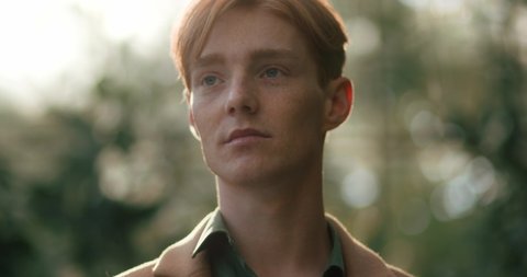 Portrait of Handsome Redhead Man opens Blue Eyes standing outdoors. Enjoys Nature at Park. Attractive Young Male with Freckles Thoughtfully looking, wears Coat and Shirt. Caucasian Ginger Guy.