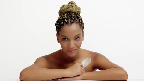 Skincare. Black Millennial Woman With Braided Hairstyle Stroking Neckline With Feather Looking At Camera Posing Over White Background In Studio. Body And Skin Care Concept. Slowmo