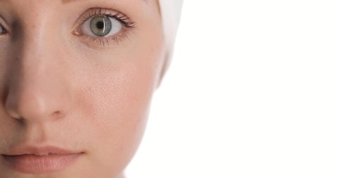 Close-up of a part of european young woman's face with a white towel on her head applies skincare cream on cheek by her hand tapping, smiling for the camera on white background. Skin care concept.