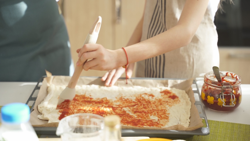 Cropped female hand adding sause on pizza base, spreading tomato sauce, preparing for baking, preparing tasty pizza together. At home, in kitchen. domestic atmosphere