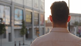 View from behind unrecognizable brunette man back view businessman guy in formal shirt tourist standing in city outdoors looking at urban buildings enjoying sunset sunlight male silhouette in sun rays