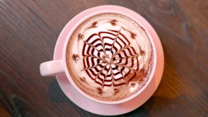 Top View of Hot Coffee Cappuccino Latte Art from Chocolate Syrup Standing on Wooden Table in Cafe. Cappuccino with Beautiful Foam in Pink Cup in Restaurant with Flower and Hearts Design on Milk Froth | Shutterstock HD Video #1080888776