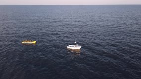 Aerial footage of a speedboat towing an inflatable banana boat at high speed. Video was shot from a drone pursuing the boat. Joyriding at sea side, nautical fun. 