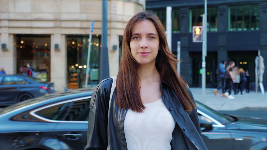 Confident businesswoman walking down street, smiling, looking at camera. Beautiful career girl in leather jacket going around city. Slender long-haired woman in hurry on business. Entrepreneuse lady