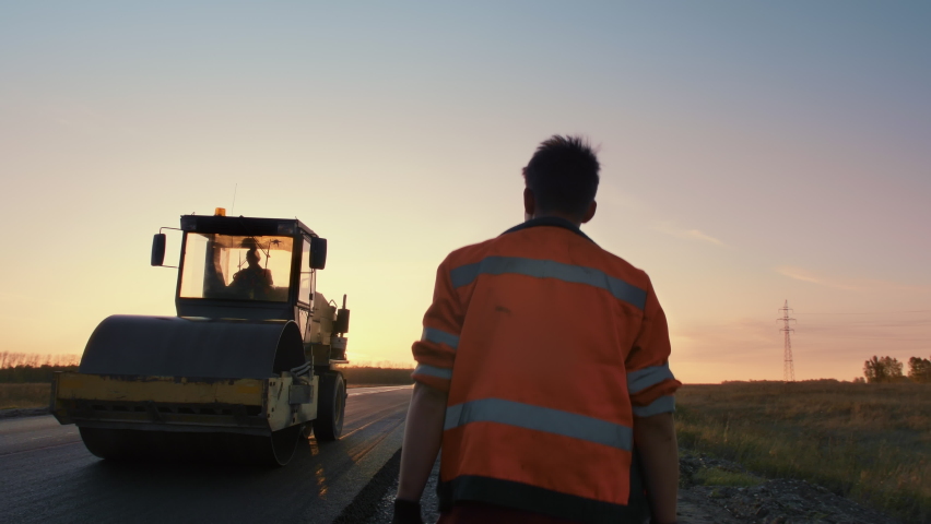 A male road worker in uniform walks to a road roller in the rays of the setting sun. Shooting from the back. Construction of a new road.  | Shutterstock HD Video #1080889853