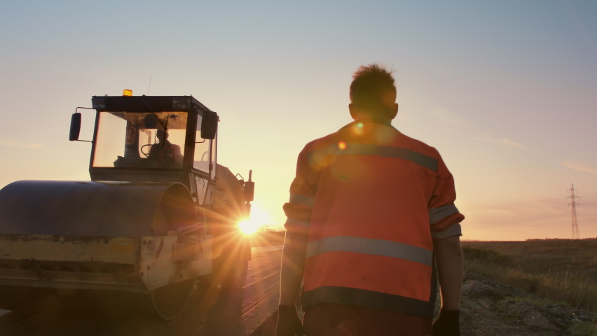 A male road worker in uniform walks to a road roller in the rays of the setting sun. Shooting from the back. Construction of a new road. 
