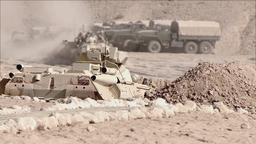 A column of armored vehicles with soldiers on armor drive past military equipment in the desert, selective focus. Concept: a combat group in a military conflict zone.