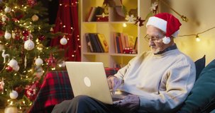 Old man pensioner speaking on video call using laptop computer. Grandfather talking online and smiling, sitting on sofa in decorated living room. Christmas holidays during coronavirus lockdown.