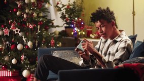 African American man playing video game on smartphone, sitting on sofa. Black afro guy gaming online, winning match. Christmas time concept. New Year holidays. Cozy winter evening at home. 