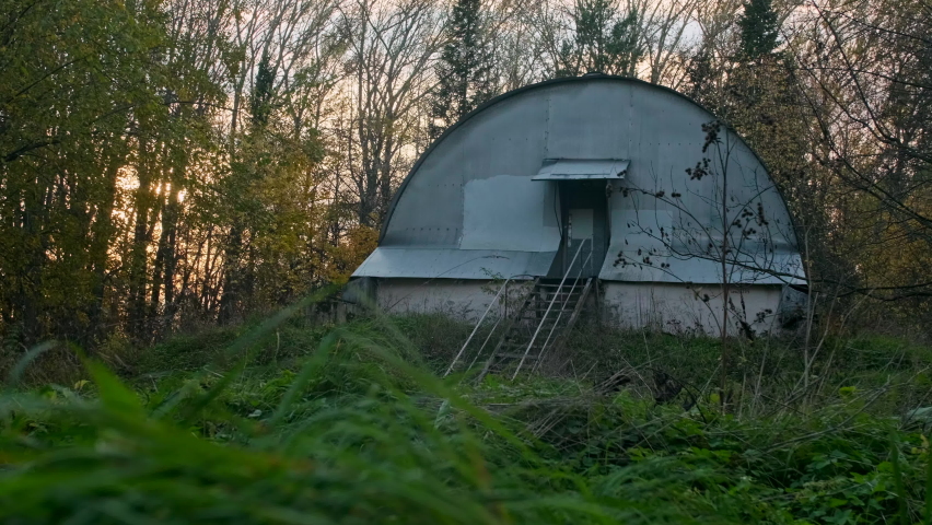 A wierd hangar or old shed with the semi-sphere roof and a metal ladder seems to be in the middle of nowhere. Industrial premises somewhere in a factory area among usual trees and coniferous trees.
