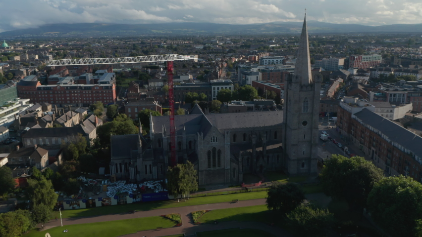 Slide and pan footage of old stone church surrounding park and town in background. Red and white tower crane. Dublin, Ireland