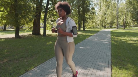 Full-length slowmo shot of young plus-size African-American woman in sportswear setting up timer on her smartwatch while jogging in park on sunny day