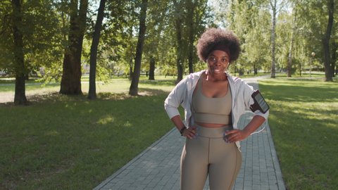 Medium slowmo portrait of smiling young African-American woman in sportswear and armband with smartphone posing for camera with hands folded standing outdoors in park during sports training