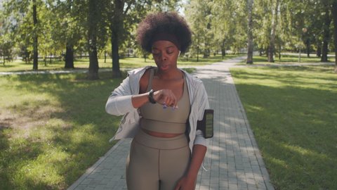 Stab slowmo shot of active young plus-size African-American woman in tight sportswear finishing jogging in park on sunny day, looking at timer on her smartwatch, smiling because of good result