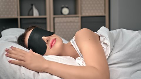 Closeup happy brunette woman in black sleep mask relaxing in comfortable bed hugging white cotton pillow. Smiling female resting enjoy dream with closed eyes under comfy blanket with positive emotion