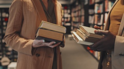 Midsection slowmo shot of hands of unrecognizable intelligent multi-ethnic couple in smart casualwear holding small pile of books at library during conversation