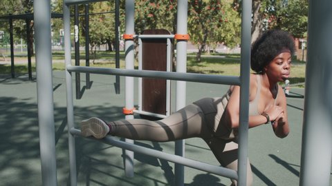 Slowmo stab shot of young plus-size Black woman in tight sportswear doing stretching exercises while training outdoors at sports ground with gym equipment