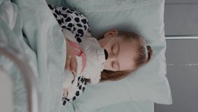 vertical video: Portrait of tired sick child sleeping after suffering medical recovery surgery during disease examination in hospital ward. Hospitalized kid resting in bed wearing oxygen nasal tube