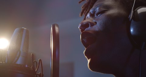 Cinematic macro shot of young professional african male singer is performing a new song with a microphone while recording it in a music studio with manager.