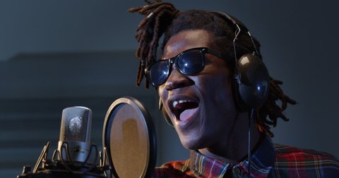 Cinematic close up shot of young professional african male singer is performing a new song with a microphone while recording it in a music studio with manager.