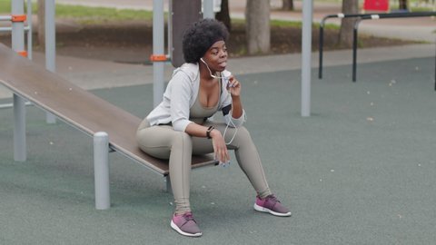 Stab slowmo shot of active young African-American woman talking through microphone on earphone wire sitting on exercise facility at sports ground outdoors on summer day