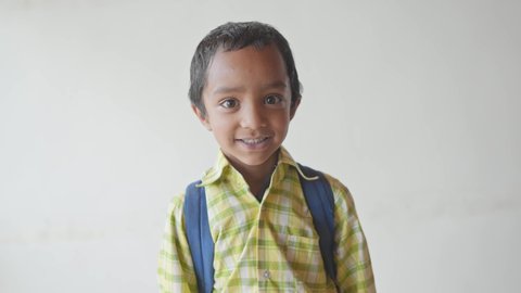 close shot of a little adorable primary school boy wearing a uniform with a backpack smiling joyfully staring at the camera standing against a white background wall. Learning and education concept