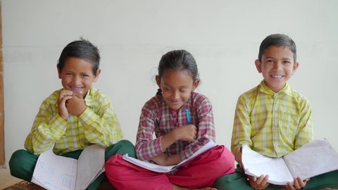 shot of trio Indian Asian primary school children wearing uniforms sitting together with text and note books looking happy against a white background or wall. learning and education concept