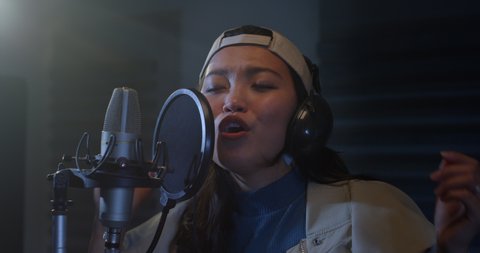 Cinematic shot of young professional energetic asian female singer is performing a new song with a microphone while recording it in a music studio with manager.