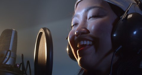 Cinematic macro shot of young professional energetic asian female singer is performing a new song with a microphone while recording it in a music studio with manager.