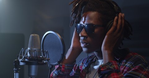 Cinematic close up shot of young professional african male singer is performing a new song with a microphone while recording it in a music studio with manager.