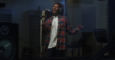 Cinematic shot of young professional african male singer is performing a new song with a microphone while recording it in a music studio with manager.